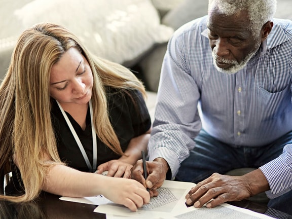 Clinician assisting an older adult with cognitive communication disorder during a writing exercise at home.