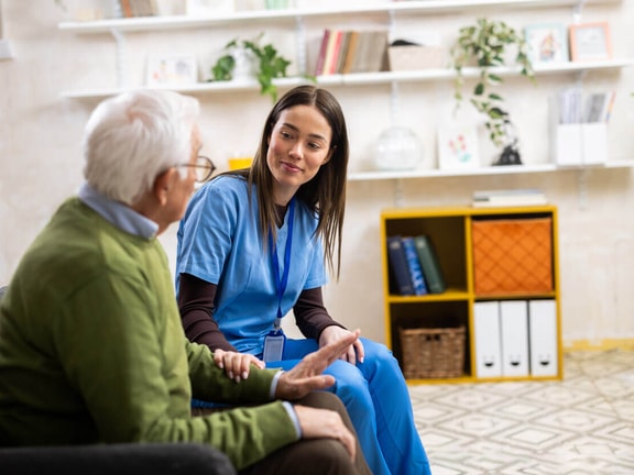 A nurse sits at eye level with an older patient, leaning in and listening during a calm bedside conversation.