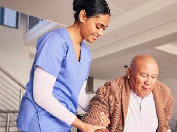 A Medbridge caregiver in blue scrubs helps an elderly man in a cozy living room.