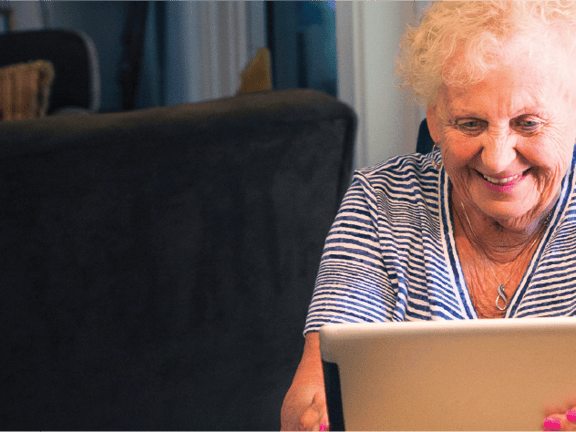 Smiling elderly woman with curly hair uses a tablet at a table with yellow flowers.