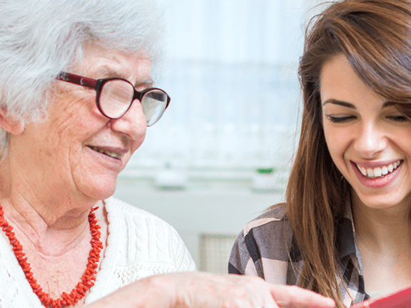 An elderly woman with glasses and a young woman with brown hair smile while reading a red book together.