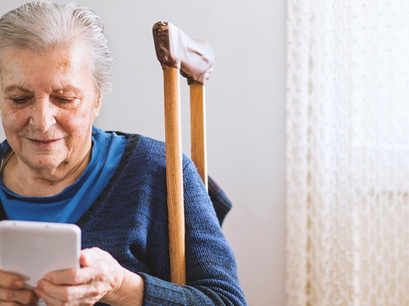 An elderly woman in a blue outfit focuses on her smartphone near lace-curtained window.
