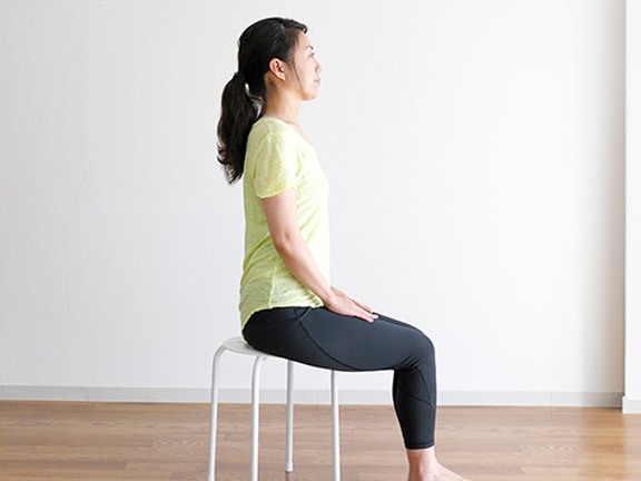 Someone in yellow and black sits on a stool in a minimalist room with a sofa, facing forward.