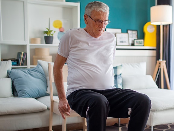 Elderly man in a white shirt and black pants sitting on a sofa in a living room with bookshelves and plants, near a large window at medbridge.
