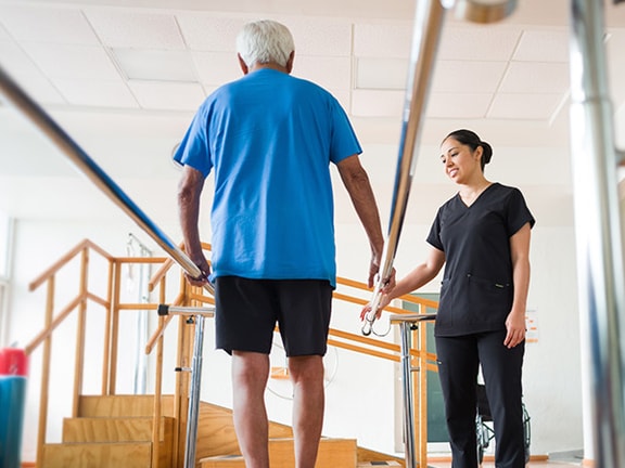 An older adult uses parallel bars to walk with help from a healthcare professional at a rehabilitation facility.