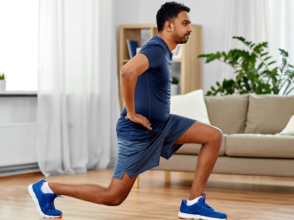 A man in athletic wear lunges in a bright living room by large windows.