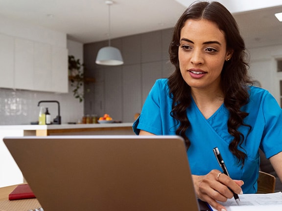 A woman in blue scrubs works at a laptop with a pen and notebook in a modern room.