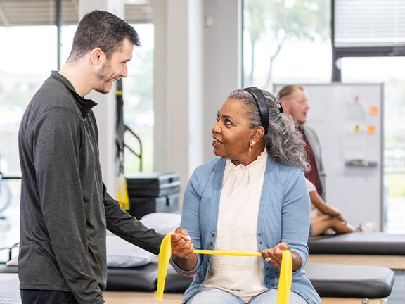 A woman on an exam table holds a yellow exercise band while talking to a man, with two others conversing nearby.