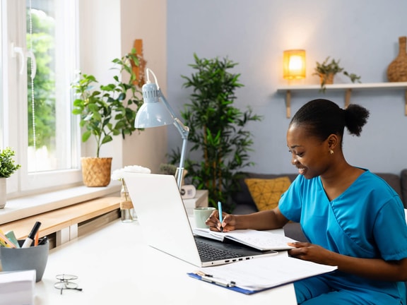 A woman in scrubs writes in a notebook at her desk while using a laptop, in a bright, modern medbridge workspace.