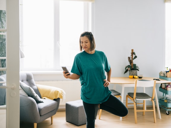 A woman in a teal shirt balances on one leg while looking at her phone in a living room near furniture.