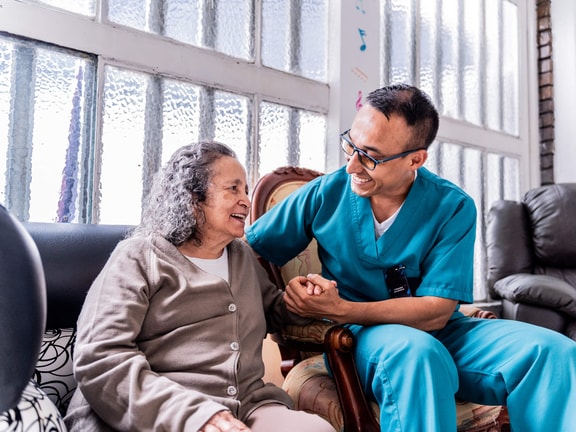 A male healthcare professional wearing teal scrubs and glasses sits next to an elderly woman, smiling warmly and holding her hand in a brightly lit room.