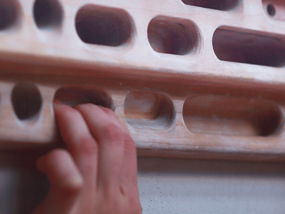 Hands holding a wooden hangboard with slots and holds for climbing practice.