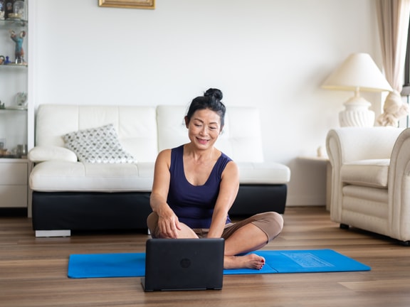 A woman smiles as she sits cross-legged on a yoga mat in her living room, looking at a medbridge course on her laptop.