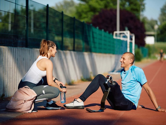 A male runner with a prosthetic blade leg sits on a track, smiling at a female coach.