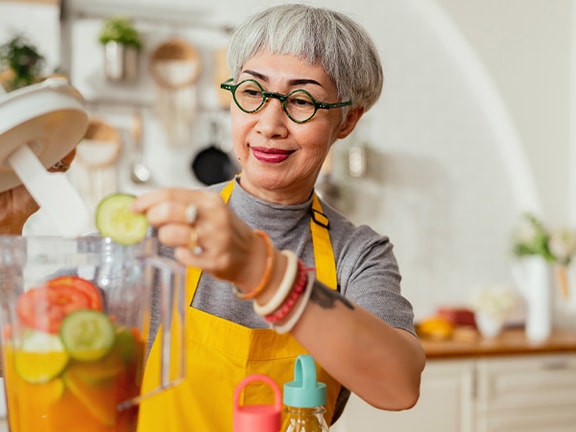 In a kitchen, someone in glasses and a yellow apron adds cucumbers to a blender.