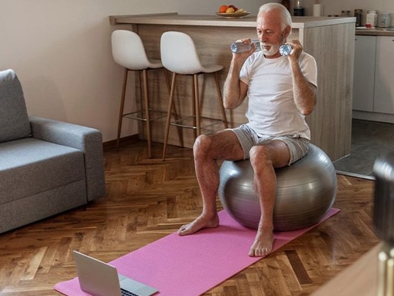 Man sitting on an exercise ball, drinking water, with a yoga mat and laptop nearby in a living room.