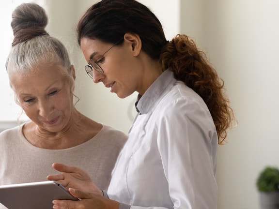 A woman in a lab coat shows a tablet to an older woman in a well-lit room, demonstrating medbridge.