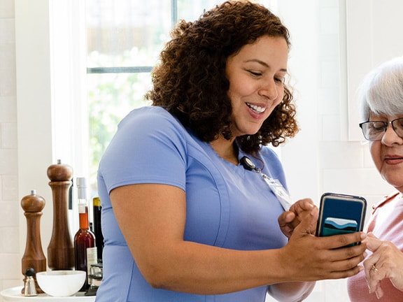 Two women in a kitchen, one sharing her smartphone screen with the other amidst cooking utensils and cutting boards; medbridge styling.