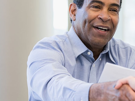 Two people are shaking hands across a table while another person sits nearby, smiling.