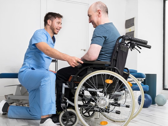 A therapist kneeling and assisting a wheelchair user with positioning in a bright, modern therapy clinic.