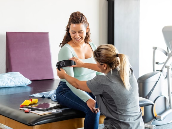 A physical therapist applies a blood flow restriction training cuff to a patient's upper arm during a rehabilitation session.