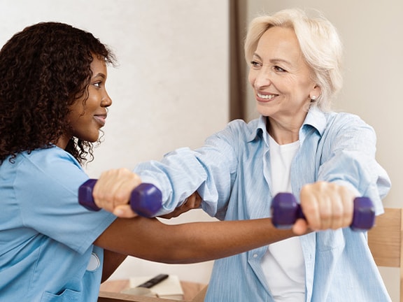 A caregiver helps an elderly woman exercise with purple dumbbells in a well-lit room.