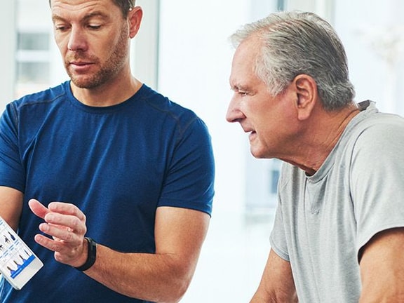 A man in a blue shirt presents a tablet to an older man in a gray shirt inside, showcasing medbridge.