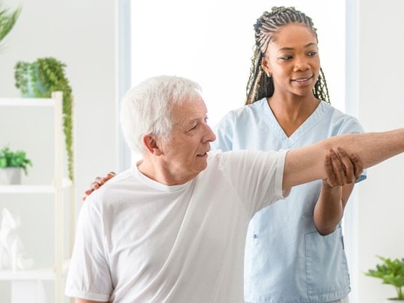 A caregiver helps an elderly man with a shoulder exercise in a bright room with anatomical charts and plants.