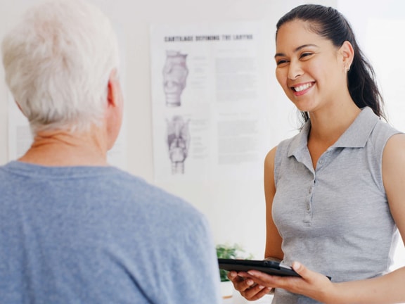 A woman with a tablet smiles at an older man in a medical office, showing the benefits of medbridge digital healthcare.