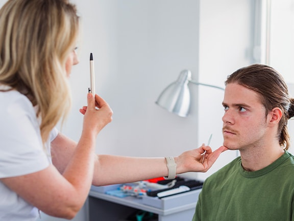 A healthcare worker uses a penlight for an eye exam on a seated person in a well-lit room.