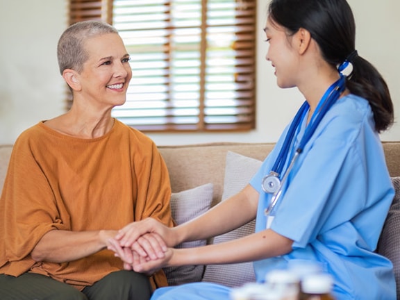 A nurse in blue scrubs chats and smiles with someone in an orange shirt on a couch.