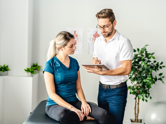 A woman sits on an exam table as a male healthcare professional takes notes, illustrating patient care using medbridge software.
