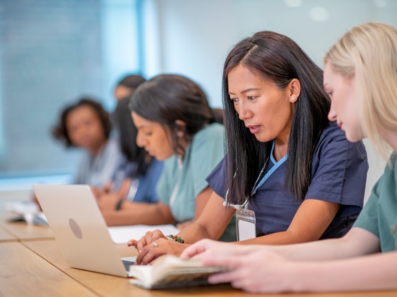 Healthcare professionals in blue and green scrubs are seated at a table, focused on learning. Two women in the foreground are using a laptop and a book. The setting is a brightly lit room. The atmosphere is professional and focused.