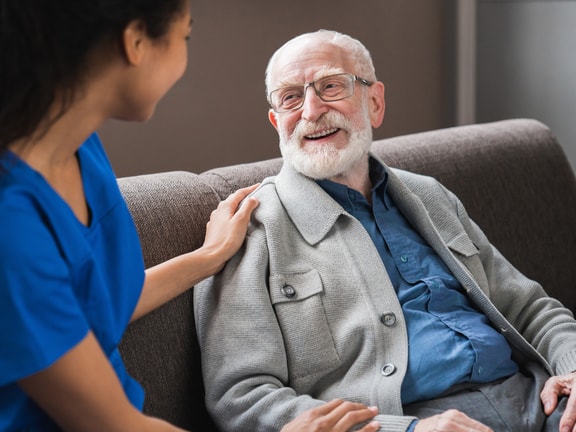 A caregiver, appearing to be a younger woman with dark hair and wearing a blue scrub top, gently places her hand on the shoulder of an older adult man with white hair and a beard, who is seated in a brown armchair and smiling. The setting appears to be indoors, possibly a care facility or the man's home. The interaction suggests a caring and supportive relationship.