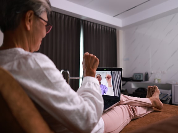 An older woman with short grey hair and red-rimmed glasses sits comfortably on a brown bed, looking at a laptop resting on her lap. She is wearing a white long-sleeved shirt and light pink pants. On the laptop screen, a younger woman in a light purple shirt smiles and holds up a closed fist in a gesture of encouragement or success. The background shows a bedroom setting with dark curtains, white walls, and a bedside table. The scene suggests a virtual connection or online interaction between the two women.