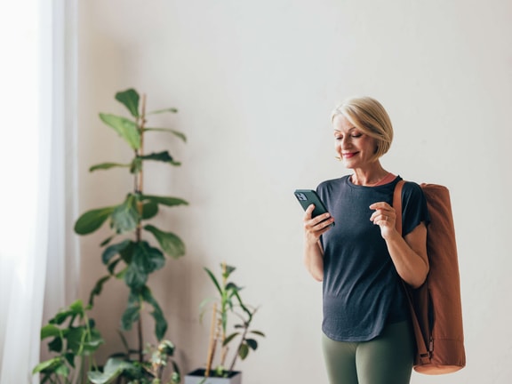 Older woman with short blonde hair and a yoga mat checks her phone indoors near potted plants, exploring digital healthcare options from medbridge.