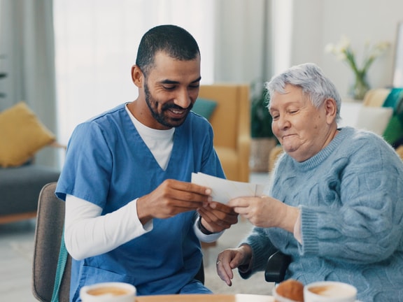A caregiver in blue scrubs assists an elderly woman with reading at a table, featuring cups and a notepad.