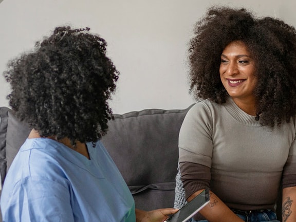 Two curly-haired individuals converse on a couch; one has a clipboard, and the other is smiling.