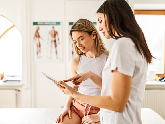 A healthcare provider shows a young woman information on a tablet in a medbridge exam room.