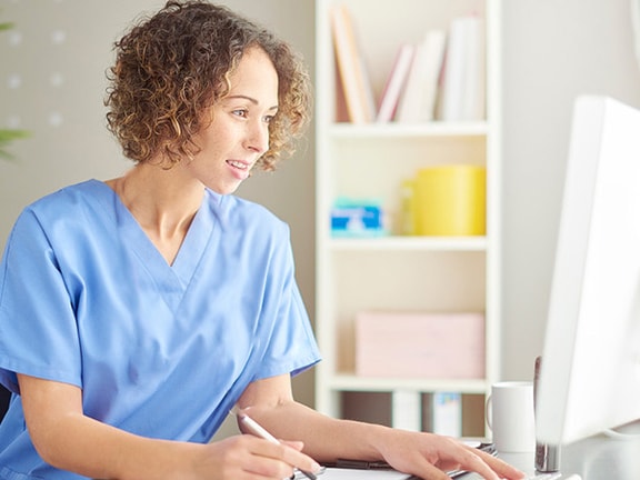 A person in blue scrubs writes at a desk with a computer, plant, and shelves visible.