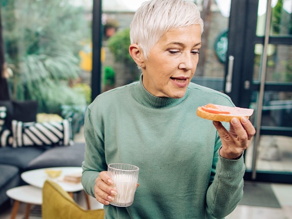 A woman with short gray hair holds a grapefruit slice and a glass of water in a living room with large windows.