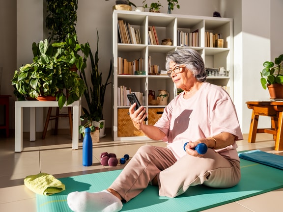 Older woman on a yoga mat holds a dumbbell and checks her phone in a bright room with medbridge training gear and plants.