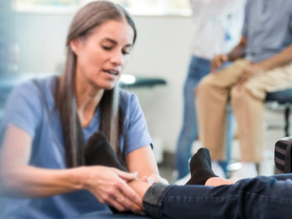 A physical therapist assesses a patient's ankle in a clinic; medbridge session visible in background.