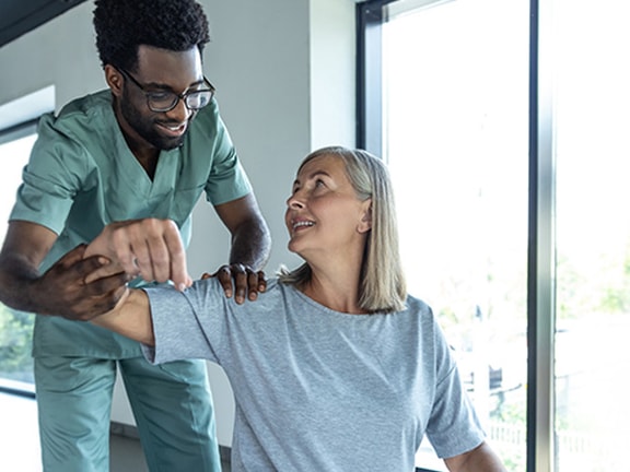 A physical therapist assisting a smiling older adult with arm exercises, demonstrating patient engagement best practices during rehabilitation for lateral elbow pain.