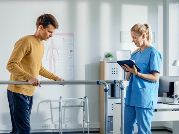 A man in a mustard shirt uses parallel bars as a woman in blue scrubs takes notes at a Medbridge clinic.