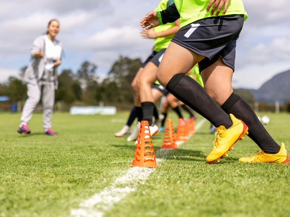 Soccer player executing cone drills on a field under coach supervision for sports injury prevention.