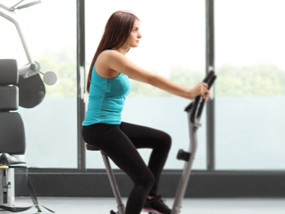 A woman works out on a stationary bike at medbridge gym with equipment in the background.