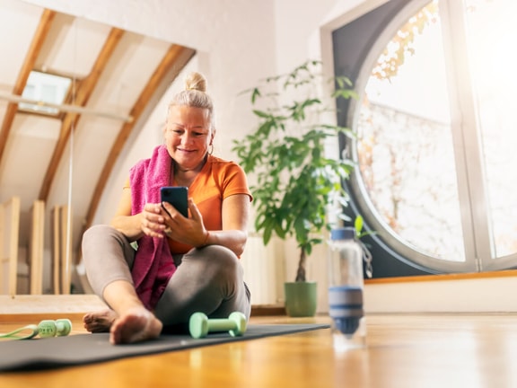 Older woman on a yoga mat smiles at her phone while using medbridge, with dumbbells and a water bottle nearby.