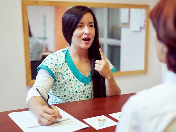 A woman in scrubs gestures while speaking to another woman at a Medbridge office desk.