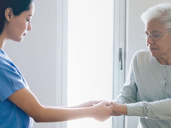 A medbridge healthcare professional in blue scrubs aids an elderly woman by a window.